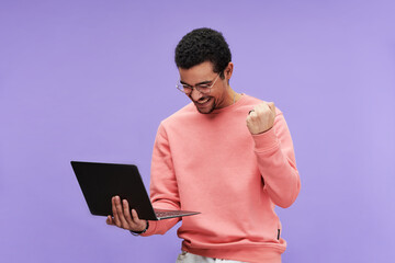Young excited programmer in eyeglasses and pink pullover expressing joy while looking at screen of laptop in his hand on violet background