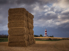 A Tower Of Straw Bales With Happisburgh Lighthouse In The Background On A Stormy Summers Evening.