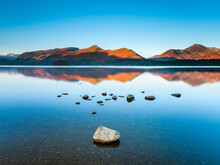 Mountains And Rocks Reflecting In The Still Waters Of Derwent Water At Sunrise.