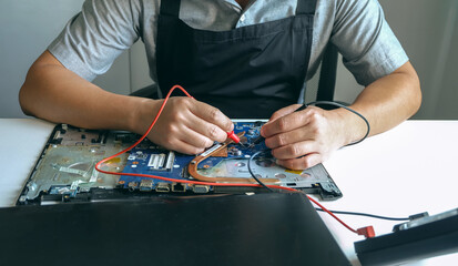 young man who is a computer technician A laptop motherboard repairman is using an IC meter to look for defects on the motherboard to repair on his desk. Board repair with modern technology	
