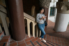 Full Length With High Angle View Of Leisure Asian Chinese Woman Visitor Standing On The Inner Spiral Staircase Of Historic Santa Barbara County Courthouse And Learning Its History On Phone