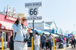 stylish asian Korean lady photographer on vacation holding hat and looking at guide on smartphone on background of route 66 end of trail sign at santa monica pier