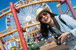 © PR Image Factory - dutch angle view of smiling asian korean lady tourist wearing sunglasses and camera on vacation at amusement park with joyful rides behind her