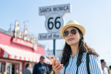 Cheerful Asian Taiwanese Lady Wearing Sunglasses Using Online Map On Phone Near Route 66 End Of The Trail Sign While Touring Santa Monica Pier In California Usa
