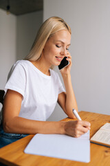 Vertical shot of pretty blonde young woman making phone call, taking notes, businesswoman consulting client or discussing project with colleague, sitting at table, freelancer working at home.