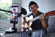 Asian influencer playing guitar during podcast or live video broadcast for the audience from the mobile phone at home