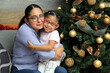 © Arlette - Divorced single mom and daughter Latina brunettes with glasses have Christmas presents sitting by the tree with spheres give each other hugs and kisses show their love in solitude