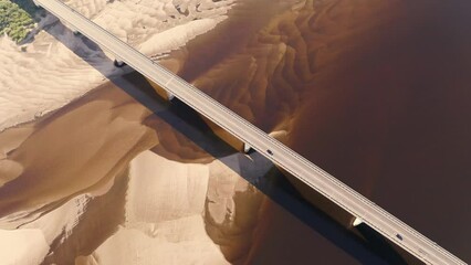 Wall Mural - Aerial drone shot of cars driving on bridge over river and sandy riverside at sunset. Amazing aesthetic 4k top view shot with beautiful coast with dune sands. Drone aerial bridge with car traffic