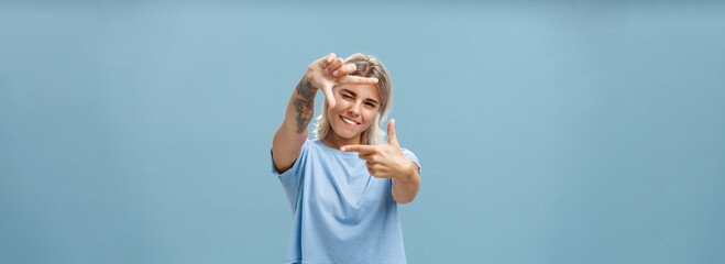 Be creative and happy. Portrait of charming enthusiastic female designer with blond hair and tattoo on arm winking and smiling joyfully making frame gesture as if measuring space over blue background