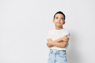 Wall Mural - a cute, pensive boy, of school age, is standing in a white T-shirt on a light background with an empty space for inserting an advertising layout with his arms crossed on his chest