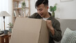 © PRPicturesProduction - cheerful asian young male is looking into box at his online shop order on the sofa at home.