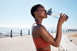 Black woman, drinking water or bottle in fitness workout, training or exercise by beach, ocean or sea in summer location. Smile, happy or sports runner with drink for healthcare wellness recovery