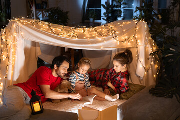 Happy family, mother, father and daughter lying inside self-made hut, tent in room in the evening and reading book. Winter coziness