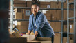 © Gorodenkoff - Portrait of a Happy Black Small Business Owner Working on Laptop in Warehouse, Preparing Parcels for Delivery. Female Smile and Pose for Camera in Storeroom with Cardboard Boxes.