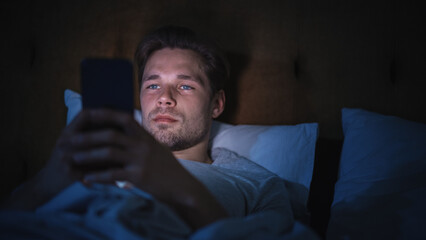 Leinwandbilder - Caucasian Man Uses Smartphone in Bed at Home at Night. Handsome Guy Browsing Social Media, Reading News, Doing Online Shopping, Chatting with Friends Late at Night.