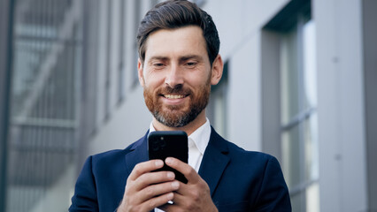 Smiling happy man holds phone browsing social network standing outdoors successful businessman responding to business or friendly email message male client makes order in online store using smartphone