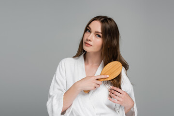 young woman brushing shiny and healthy hair while looking at camera isolated on grey