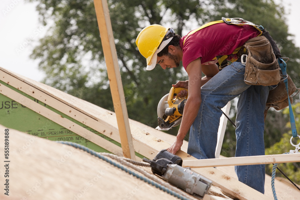 Hispanic carpenter using a circular saw on the roof at a house under ...