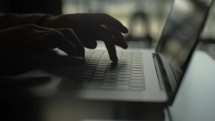Close-up side view of unrecognizable male programmer freelancer working typing on keyboard laptop computer sitting at desk in dark home office room, on background of window. Shooting in slow motion.