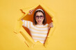 Portrait of positive optimistic woman wearing striped T-shirt, hair band and sunglasses, breaking through paper hole in yellow wall, looking at camera, showing tongue out.