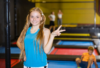 Cheerful confident sporty teen girl enjoying time in indoor trampoline arena, standing with jumping children in background and giving V sign