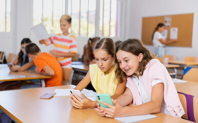 Girls sitting at desk in classroom, using smartphones together during lesson in school.