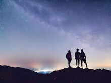 Silhouette Of Young Couple Hiker Were Standing At The Top Of The Mountain Looking At The Stars And Milky Way Over The Twilight Sky. Both Of Them Were Happy And Free To Travel