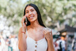 © dzmatph - Happy Latin American female tourist with backpack smiling and looking away with smile while talking on cellphone on blurred background of sunlit park in Barcelona, Spain