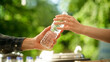 © Gorodenkoff - Close Up of a Anonymous Person Handing Over a Water Bottle to Another Person. Green Background in Nature. Outdoors Fourt Court Selling Drinks. Ecology, Healthcare and Hydration Concept.
