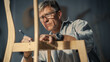© Gorodenkoff - Close Up of a Carpenter Putting on Glasses, Looking at a Blueprint and Starting to Assemble a Wooden Chair. Professional Furniture Designer Working in a Studio in Loft Space with Tools on the Walls.
