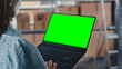 © Gorodenkoff - Woman Using a Laptop Computer with Green Screen Chromakey Mock Up Display. Employee Browsing Internet, Using Trackpad in an Online Store Warehouse Storeroom with Parcels. Over the Shoulder Shot.