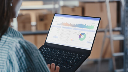 Woman Using a Laptop Computer with Dashboard, Analysis Charts Display. Employee Browsing Internet, Using Trackpad in an Online Store Warehouse Storeroom with Parcels. Over the Shoulder Shot.