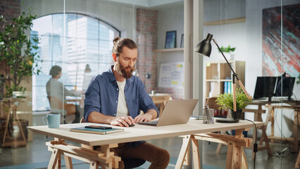 Fototapete - Stylish Long-Haired Bearded Specialist Sitting at a Desk in Creative Agency. Young Stylish Man Working on a Laptop Computer in Marketing Company. Office Team Members in the Background.