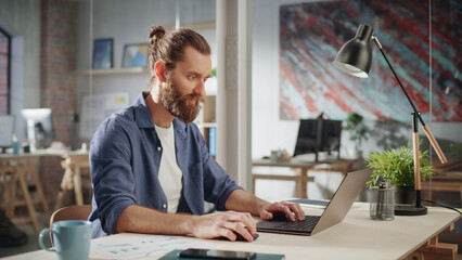 Wall Mural - Handsome Long-Haired Bearded Manager Sitting at a Desk in Creative Office. Young Stylish Man Using Laptop Computer in Marketing Agency. Colleagues Working in the Background.