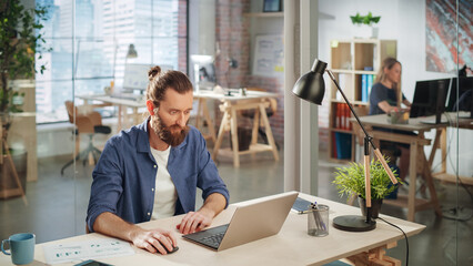 Fototapete - Stylish Long-Haired Bearded Specialist Sitting at a Desk in Creative Agency. Young Stylish Man Working on a Laptop Computer in Marketing Company. Office Team Members in the Background.