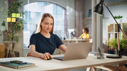Fototapete - Beautiful Female in a Black Casual Dress Sitting at a Desk in Creative Office, Working on Tasks on Laptop Computer. Smiling Creative Specialist Writing Corporate Project Plans for Marketing Agenda.