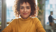 Portrait Of A Beautiful Middle Eastern Manager Sitting At A Desk In Creative Office. Young Stylish Female With Curly Hair Looking At Camera With Big Smile. Colleagues Working In The Background.