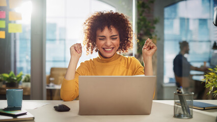 Leinwandbilder - Portrait of a Beautiful Middle Eastern Manager Sitting at a Desk in Creative Office. Young Stylish Female with Curly Hair Celebrating her Achievement with Big Smile