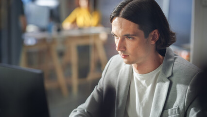 Poster - Close Up Portrait of a Handsome Manager Sitting at a Desk in Creative Office. Young Stylish Man in a Jacket Working on a Computer in Marketing Agency. Colleagues at Work in the Background.