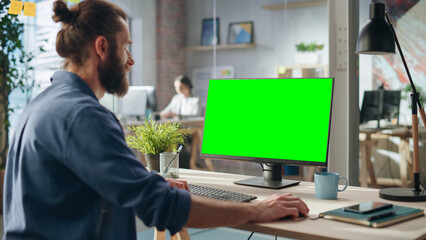 Wall Mural - Handsome Long-Haired Bearded Manager Working at a Desk in Creative Office, Using Desktop Computer with Green Screen Mock Up Display. Colleagues Working in the Background in Marketing Agency.