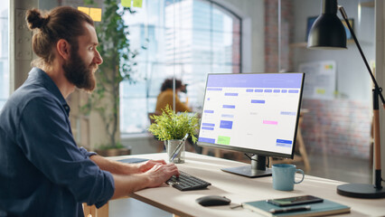 Aufkleber - Handsome Long-Haired Bearded Manager Working at a Desk in Creative Office, Using Desktop Computer with a Calendar Software on Screen. Colleagues Working in the Background in Marketing Agency.
