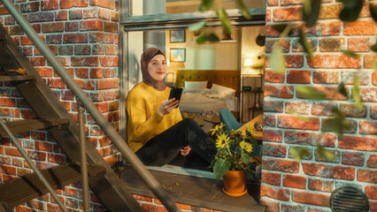 Poster - Positive, Smiling Muslim Female Using a Smartphone while Sitting on a Window Sill in Cozy Brownstone House Apartment. Girl Checking Online Social Media. Tilt Down Shot from Outside the Building.