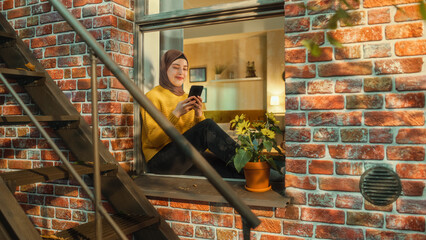 Poster - Young Arab Woman Using a Smartphone while Sitting on a Window Sill in a Multi-Storey Brick Apartment Building. Multiethnic Female Connecting with Friends Online. Tilt Up Shot from Outside the House.