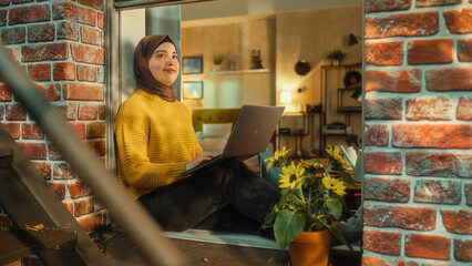 Poster - Portrait of a Young Muslim Female in Hijab Working on a Laptop Computer while Sitting on a Window Sill. Business Office Employee Working Remotely from Home. View from Outside the Window.