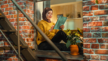 Poster - Happy Arab Female in a Hijab Sitting on a Window Sill, Checking Social Media, Using Online Apps on Tablet Computer. Shot from Apartment Window. Brownstone Multi-Storey Building with Emergency Stairs.