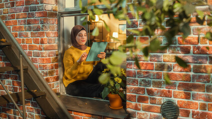 Poster - Portrait of a Young Arab Woman Using Tablet Computer while Sitting on Window Sill in a Multi-Storey Brick House. Multiethnic Female Connecting with Friends Online. Shot from Outside the House.
