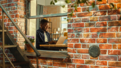 Poster - Young Handsome Male Using Laptop Computer, Sitting in Living Room in Apartment. African American Businessman Checking Social Media, Smiling, Answering Emails from Home. Inside Apartment Window View.