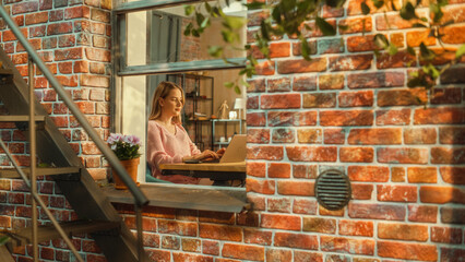 Poster - Young Attractive Female in Pink Sweater Working on a Laptop Computer while Sitting Behind a Table in Living Room. Business Office Employee Working Remotely from Home. View from Outside the Window.