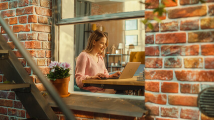 Poster - Portrait of a Young Female Student Using Laptop Computer while Sitting Behind a Table in Her Stylish Living Room. Successful College Graduate Studying Remotely from Home. Shot from Outside the Window.