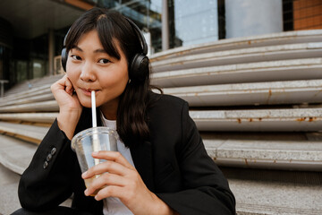 Wall Mural - Young asian woman in headphones smiling and drinking juice outdoors